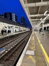A train platform with tracks running parallel, bordered by a covered station with white beams. Several people are walking along the platform, and tall buildings can be seen in the background against a dark blue evening sky. The platform is illuminated by overhead lights, creating a contrast with the dark surroundings.