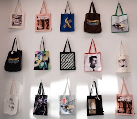 A collection of tote bags, each with a unique design, hangs on a white wall. The designs include art prints, graphic texts, and images of people. The bags are arranged in a grid with varied colors and artistic themes.