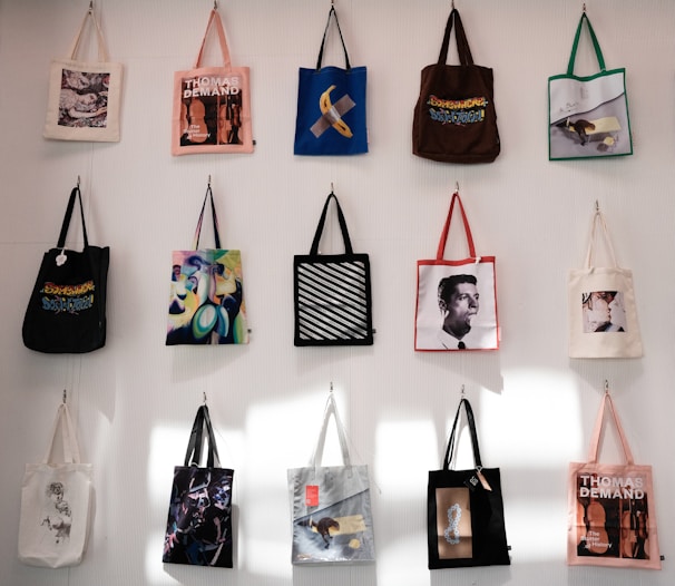 A collection of tote bags, each with a unique design, hangs on a white wall. The designs include art prints, graphic texts, and images of people. The bags are arranged in a grid with varied colors and artistic themes.
