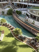 A multi-level shopping complex features a landscaped area with lush green plants and a flowing waterway. A modern bridge crosses over the water, and people can be seen walking both on the bridge and on the upper level where various shops are visible. Sunlight casts shadows providing a warm and inviting atmosphere.