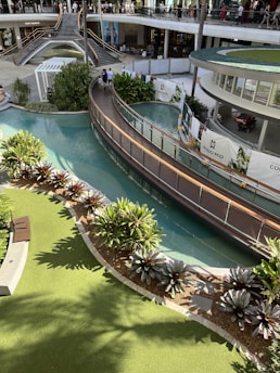 A multi-level shopping complex features a landscaped area with lush green plants and a flowing waterway. A modern bridge crosses over the water, and people can be seen walking both on the bridge and on the upper level where various shops are visible. Sunlight casts shadows providing a warm and inviting atmosphere.