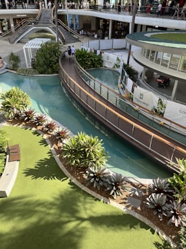 A multi-level shopping complex features a landscaped area with lush green plants and a flowing waterway. A modern bridge crosses over the water, and people can be seen walking both on the bridge and on the upper level where various shops are visible. Sunlight casts shadows providing a warm and inviting atmosphere.