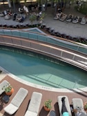 A modern outdoor relaxation area featuring a small pool with clear water, surrounded by wooden decking. Several people are lounging on cushioned chairs, enjoying the sunny weather. Lush plants and a curved wooden bridge add to the tranquil ambiance. In the background, more people are seated, creating a leisurely atmosphere.