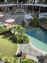 An outdoor shopping area with a modern architectural design featuring a two-level open space. The upper level has a storefront with large windows and is accessed by wide staircases flanking a waterfall. The lower level features a well-landscaped area with lush greenery and a small artificial pond. People are casually strolling and sitting around, with some seated under umbrellas. The area is bathed in sunlight, creating a vibrant atmosphere.