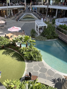 An outdoor shopping area with a modern architectural design featuring a two-level open space. The upper level has a storefront with large windows and is accessed by wide staircases flanking a waterfall. The lower level features a well-landscaped area with lush greenery and a small artificial pond. People are casually strolling and sitting around, with some seated under umbrellas. The area is bathed in sunlight, creating a vibrant atmosphere.