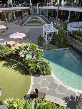 An outdoor shopping area with a modern architectural design featuring a two-level open space. The upper level has a storefront with large windows and is accessed by wide staircases flanking a waterfall. The lower level features a well-landscaped area with lush greenery and a small artificial pond. People are casually strolling and sitting around, with some seated under umbrellas. The area is bathed in sunlight, creating a vibrant atmosphere.