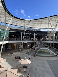 A shopping center with multiple levels featuring a modern design and a partially open roof structure. Several shops with glass fronts are visible, including well-known brands. People are walking and sitting around, and there's a fountain surrounded by a staircase. Large umbrellas provide shaded seating areas in the open plaza.