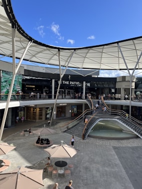 A shopping center with multiple levels featuring a modern design and a partially open roof structure. Several shops with glass fronts are visible, including well-known brands. People are walking and sitting around, and there's a fountain surrounded by a staircase. Large umbrellas provide shaded seating areas in the open plaza.