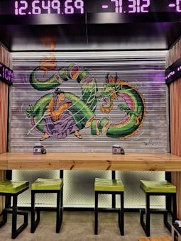 A vibrant mural featuring a green Chinese dragon intertwined with a character in traditional attire is painted on a metal roller shutter. Above, digital ticker displays show numbers in bright purple. Below the mural, a wooden table with green stools is set against a black mesh background.