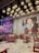 A modern, stylish restaurant interior featuring a mix of Asian cultural elements. The ceiling is adorned with a cluster of hanging, cylindrical lights, providing warm illumination. On the walls, there are large, vibrant murals, one with pink neon text saying 'THE SUSHI WAY IS MY WAY' and another depicting a woman's face with the text 'THAI LOUNGE'. Red wooden chairs surround small round tables, all set on a patterned tile floor.