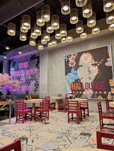 A modern, stylish restaurant interior featuring a mix of Asian cultural elements. The ceiling is adorned with a cluster of hanging, cylindrical lights, providing warm illumination. On the walls, there are large, vibrant murals, one with pink neon text saying 'THE SUSHI WAY IS MY WAY' and another depicting a woman's face with the text 'THAI LOUNGE'. Red wooden chairs surround small round tables, all set on a patterned tile floor.