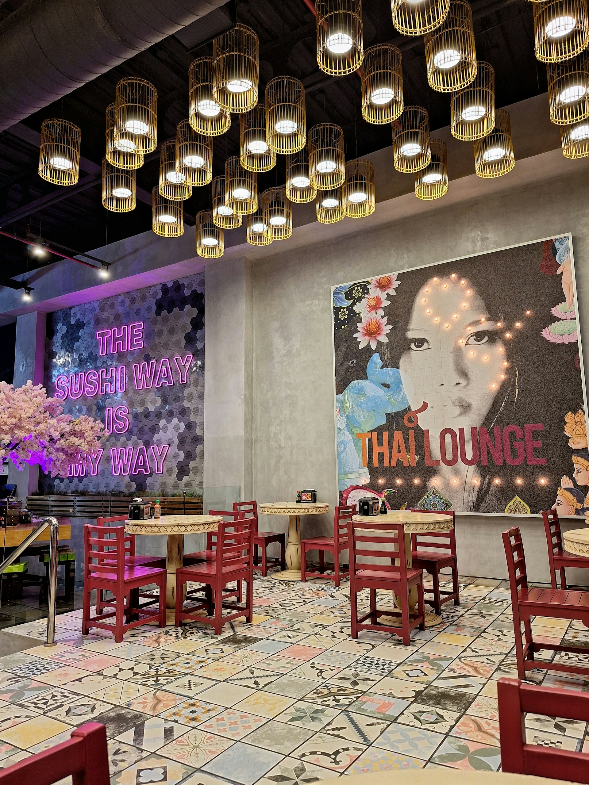 A modern, stylish restaurant interior featuring a mix of Asian cultural elements. The ceiling is adorned with a cluster of hanging, cylindrical lights, providing warm illumination. On the walls, there are large, vibrant murals, one with pink neon text saying 'THE SUSHI WAY IS MY WAY' and another depicting a woman's face with the text 'THAI LOUNGE'. Red wooden chairs surround small round tables, all set on a patterned tile floor.