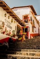 A rustic and colorful outdoor scene featuring a charming street with stone steps leading up to a quaint restaurant with a sign reading 'Restaurante Hosteria-Bar Adobe'. The building is adorned with wooden roofs, hanging lanterns, red flowers, and decorative objects such as reindeer figures. Two people are seated on the steps, adding a human element to the lively ambiance.