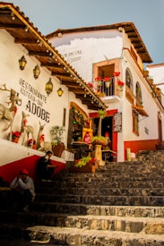 A rustic and colorful outdoor scene featuring a charming street with stone steps leading up to a quaint restaurant with a sign reading 'Restaurante Hosteria-Bar Adobe'. The building is adorned with wooden roofs, hanging lanterns, red flowers, and decorative objects such as reindeer figures. Two people are seated on the steps, adding a human element to the lively ambiance.