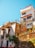 A colorful hillside house with multiple levels featuring vibrant architectural details and potted plants. The building has a rustic charm with its mix of white walls, exposed brick, and decorative ironwork. Bright flowers and a clear blue sky add to the lively and vibrant atmosphere.