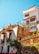 A colorful hillside house with multiple levels featuring vibrant architectural details and potted plants. The building has a rustic charm with its mix of white walls, exposed brick, and decorative ironwork. Bright flowers and a clear blue sky add to the lively and vibrant atmosphere.