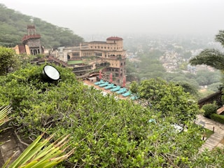 A scenic view of a large historic fort or palace surrounded by lush greenery. The structure has multiple levels, featuring arches and turrets, and is built in a traditional style with earthy tones. In the foreground, vibrant green foliage covers the landscape while a pool area with turquoise sun loungers and red umbrellas can be seen. The backdrop consists of misty hills and a distant view of a town at the bottom.