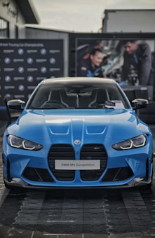 A blue BMW M4 Competition is prominently displayed, capturing the sleek and aggressive design of the car. The front grille and headlights are distinct, complementing the aerodynamic curves of the vehicle. In the background, there are blurred images of people working on a car, adding an automotive workshop ambiance.