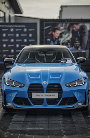 A blue BMW M4 Competition is prominently displayed, capturing the sleek and aggressive design of the car. The front grille and headlights are distinct, complementing the aerodynamic curves of the vehicle. In the background, there are blurred images of people working on a car, adding an automotive workshop ambiance.