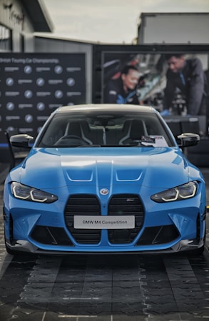 A blue BMW M4 Competition is prominently displayed, capturing the sleek and aggressive design of the car. The front grille and headlights are distinct, complementing the aerodynamic curves of the vehicle. In the background, there are blurred images of people working on a car, adding an automotive workshop ambiance.