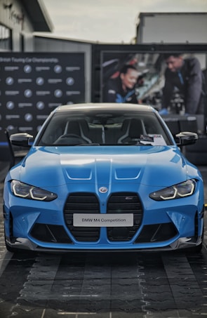 A blue BMW M4 Competition is prominently displayed, capturing the sleek and aggressive design of the car. The front grille and headlights are distinct, complementing the aerodynamic curves of the vehicle. In the background, there are blurred images of people working on a car, adding an automotive workshop ambiance.