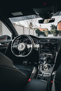 The interior of a car features a sleek black dashboard with a steering wheel bearing the Volkswagen logo. The center console includes various controls, a gear shift, and a modern infotainment system. The car seats are upholstered in dark material, and the view through the windshield shows a residential street with trees and houses in the background.