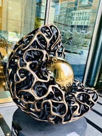 A contemporary sculpture featuring an intricate, abstract design with a metallic lattice structure enveloping a reflective golden sphere. It is placed indoors near large glass windows with reflections of a modern building, adding an urban backdrop.
