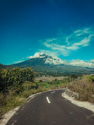 A winding road leads through lush green landscape towards a majestic mountain topped with white clouds. The sky is clear blue with scattered clouds, and greenery surrounds the path.