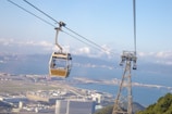 A cable car is suspended in the air over an urban landscape with a large body of water in the background. Buildings and infrastructure are visible below, as well as a green hilly area in the foreground. The sky is clear with a few scattered clouds, and distant mountains can be seen across the water.