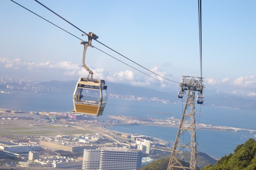 A cable car is suspended in the air over an urban landscape with a large body of water in the background. Buildings and infrastructure are visible below, as well as a green hilly area in the foreground. The sky is clear with a few scattered clouds, and distant mountains can be seen across the water.