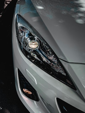 A close-up of a modern car headlight and part of the car's front body. The headlight design is sleek and features intricate light patterns, with reflective surfaces capturing surrounding light. The car appears to be parked, with shadows and subtle reflections visible on the metallic silver paint.