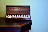 A small, wooden keyboard instrument with black and white keys sits on a wooden surface against a pale green wall. The instrument's cover is open, revealing the full keyboard.