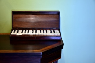 A small, wooden keyboard instrument with black and white keys sits on a wooden surface against a pale green wall. The instrument's cover is open, revealing the full keyboard.