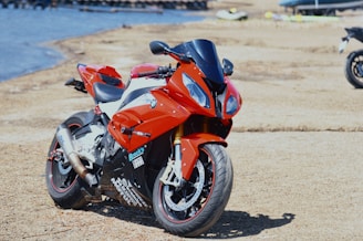 A sporty red motorcycle parked on a sandy or gravel surface near a body of water. The motorcycle features sleek lines and is equipped with visible exhaust pipes and decals.