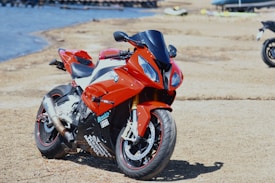 A sporty red motorcycle parked on a sandy or gravel surface near a body of water. The motorcycle features sleek lines and is equipped with visible exhaust pipes and decals.