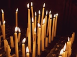 Numerous tall, wax candles are lit and arranged in rows, with soft, warm flames flickering in a dimly lit environment. Some candles are burned down more than others, and melted wax has dripped onto the surface below.