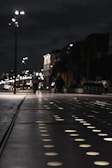 A nighttime street scene features a pathway with illuminated circular patterns. Streetlights line the path, and a few people in the distance walk along the sidewalk. Buildings with dark facades and subtle lighting can be seen, creating a modern urban atmosphere.