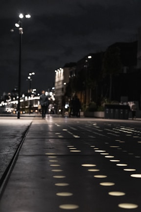 A nighttime street scene features a pathway with illuminated circular patterns. Streetlights line the path, and a few people in the distance walk along the sidewalk. Buildings with dark facades and subtle lighting can be seen, creating a modern urban atmosphere.