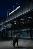 A modern building with a prominent sign reading 'Istanbul Modern' is illuminated against a dark evening sky. The architecture features sleek lines and extensive use of glass. In the foreground, two people are walking, carrying shopping bags, with the area dimly lit.