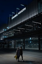 A modern building with a prominent sign reading 'Istanbul Modern' is illuminated against a dark evening sky. The architecture features sleek lines and extensive use of glass. In the foreground, two people are walking, carrying shopping bags, with the area dimly lit.