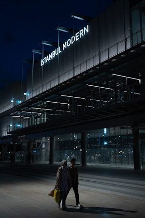 A modern building with a prominent sign reading 'Istanbul Modern' is illuminated against a dark evening sky. The architecture features sleek lines and extensive use of glass. In the foreground, two people are walking, carrying shopping bags, with the area dimly lit.