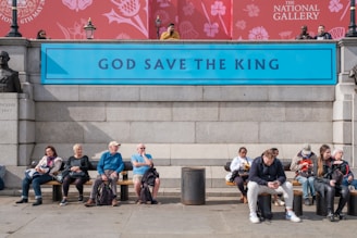 A group of people sits on a bench in front of a stone wall that features a blue sign with the text 'GOD SAVE THE KING.' The backdrop includes a red banner with floral designs and the words 'THE NATIONAL GALLERY.' A bust statue is visible on the left side of the image. Some of the individuals are engaged in eating, while others appear to be using their phones.