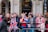 A group of people stands behind a metal barricade on a street, some holding or wearing items with the Union Jack pattern. They appear to be waiting or watching an event, and several have small British flags on headbands. The background shows a building with arches and a storefront labeled Waterstones.