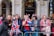 A group of people stands behind a metal barricade on a street, some holding or wearing items with the Union Jack pattern. They appear to be waiting or watching an event, and several have small British flags on headbands. The background shows a building with arches and a storefront labeled Waterstones.