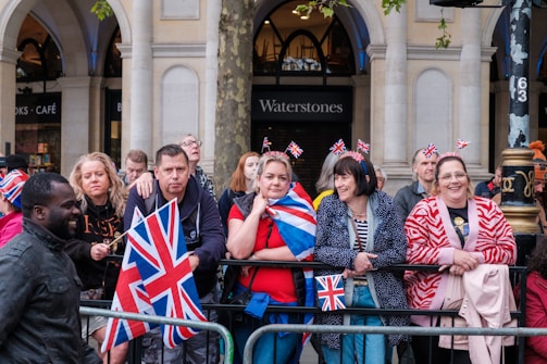 A group of people stands behind a metal barricade on a street, some holding or wearing items with the Union Jack pattern. They appear to be waiting or watching an event, and several have small British flags on headbands. The background shows a building with arches and a storefront labeled Waterstones.