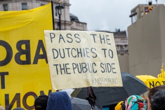 The image depicts a protest with people holding signs. One of the prominent signs reads 'PASS THE DUTCHIES TO THE PUBIC SIDE #NOTMYKING' and there is a large yellow banner in the background. People are standing together in a crowded manner, some holding umbrellas as it appears to be raining.