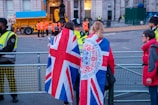 A group of people are gathered near a fenced-off area, with some wearing jackets featuring the Union Jack. Security personnel in high-visibility vests are present, and a large vehicle with a blue cover is visible in the background.