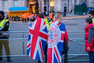 A group of people are gathered near a fenced-off area, with some wearing jackets featuring the Union Jack. Security personnel in high-visibility vests are present, and a large vehicle with a blue cover is visible in the background.