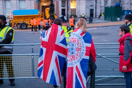A group of people are gathered near a fenced-off area, with some wearing jackets featuring the Union Jack. Security personnel in high-visibility vests are present, and a large vehicle with a blue cover is visible in the background.