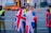 A group of people are gathered near a fenced-off area, with some wearing jackets featuring the Union Jack. Security personnel in high-visibility vests are present, and a large vehicle with a blue cover is visible in the background.